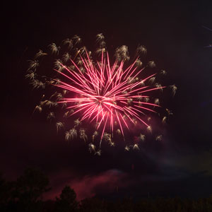 A red firework explodes in streaks in the night sky, with small yellow explosions around the edges