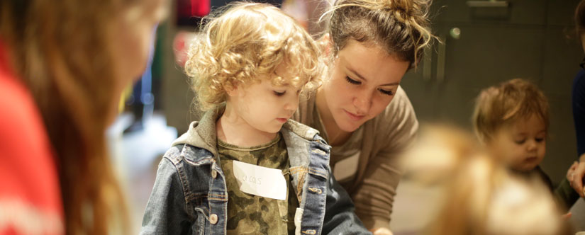 Toddler with curly blond hair is helped by mom who stand behind him