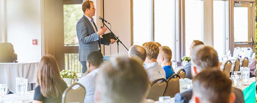 man speaking at microphone, in front of tables of crowd listening