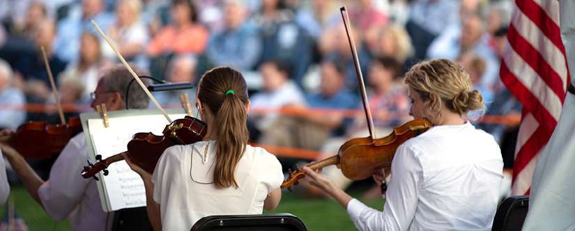 two violinists in white shirts with facing away from camera, with outdoor seated crowd in background