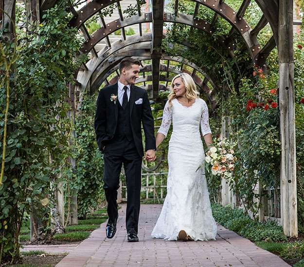 A bride and groom walk along a cobblestone path with wooden arbors above