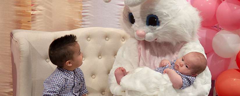 Easter Bunny mascot holds a baby and looks at a small boy seated next to him 