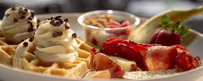 close up photo of waffles with whipped cream on top, next to bacon and potatoes in front of yogurt parfait