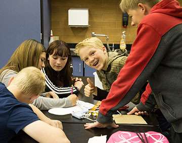 students in a classroom working around a project, a young blond boy smiles for the camera with a thumbs up