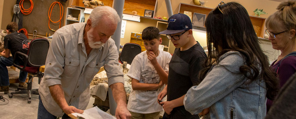 Paleontologist with white hair and button up shirt point to a clipboard for four people in front of him