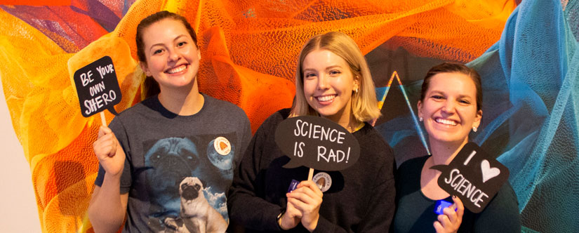 3 women in front of colorful background holding small signs that say 'Science is Rad!' and 'I Love Science'