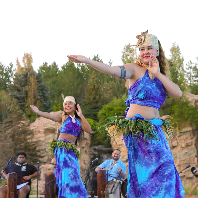 On cement pad in front of trees and rocks, 3 Polynesian women with green leaved skirts and pink and white leis dancing 
