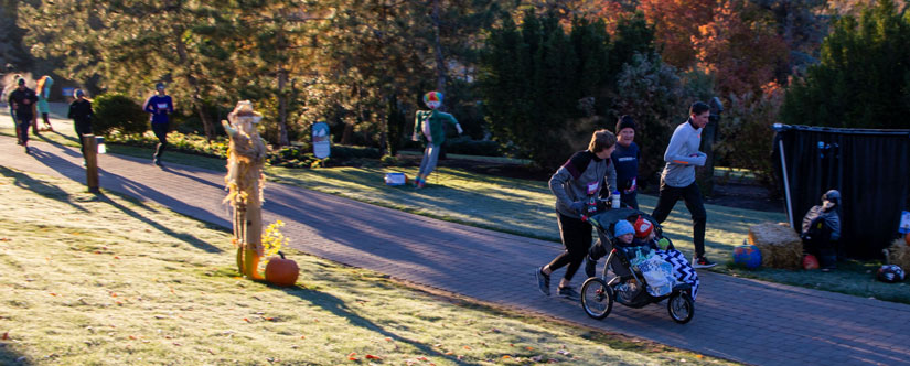 runners in beanies pushing strollers along path next to scarecrow