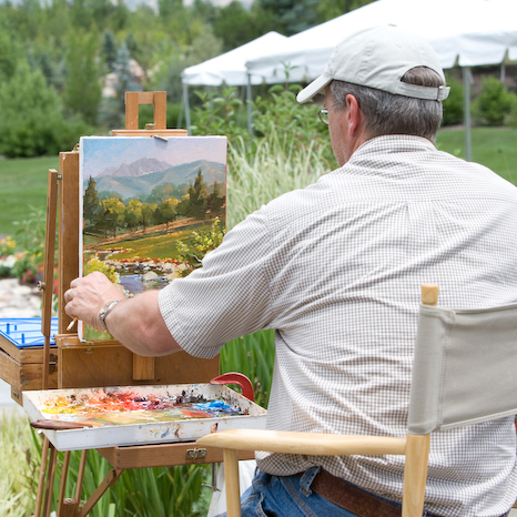 man faces away from camera outside, painting the green landscape in front of him