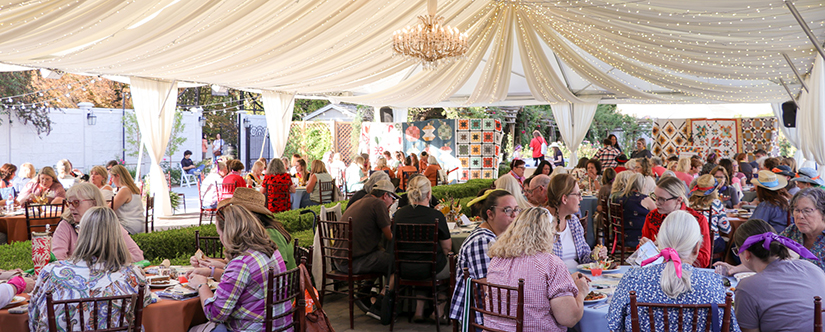 women under tent at tables surrounded by quilts