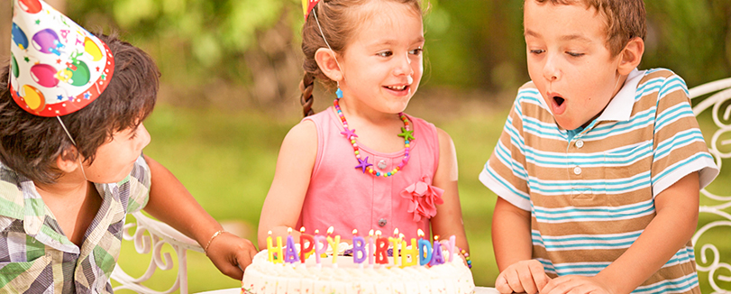 children outside, one blows out candles on cake while others watch with party hats
