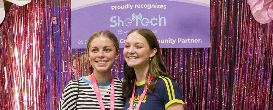 two teenage girls with pink lanyards in front of thanksgiving point SheTech sign and sparkly backdrop
