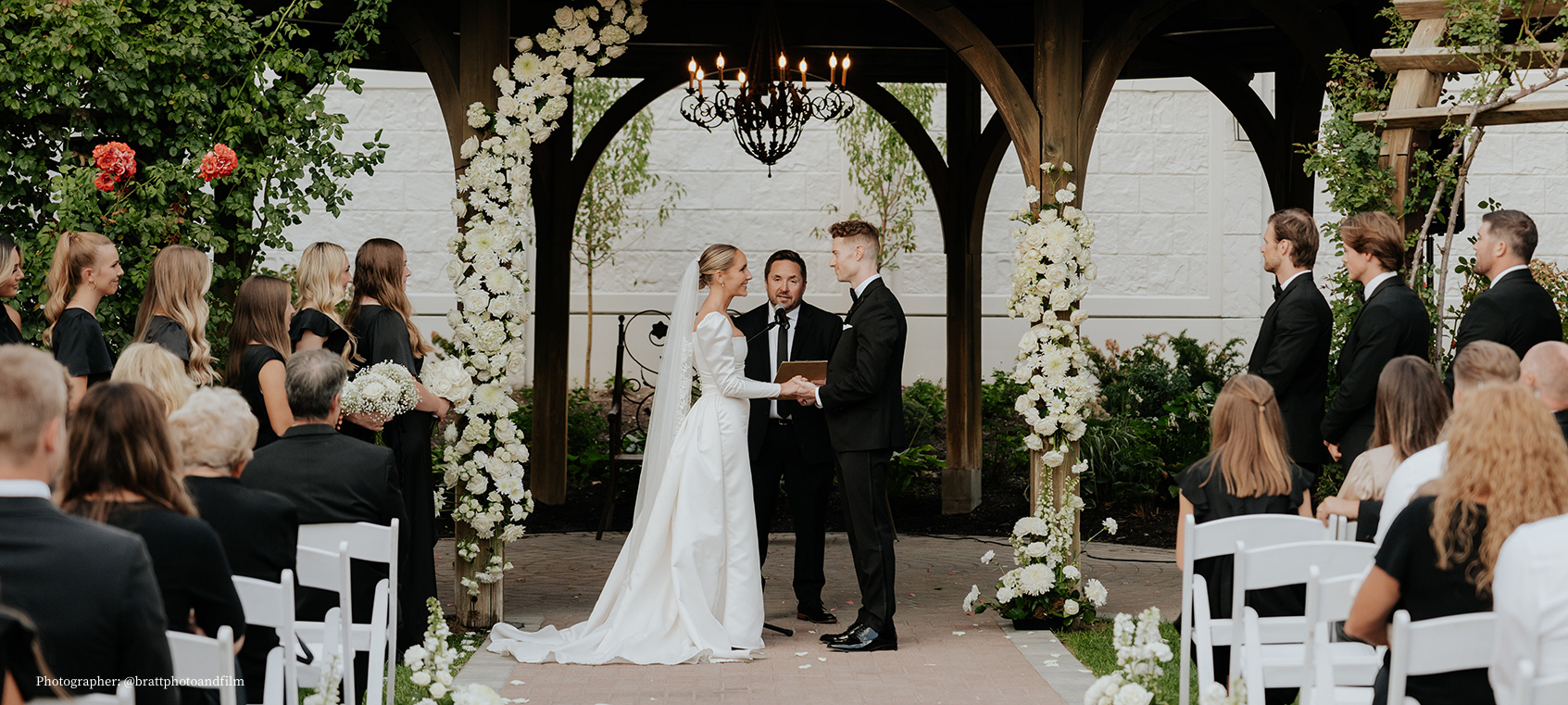 bride and groom at ceremony, chairs with guests facing them, surrounded by flowers on arch outdoors at thanksgiving point